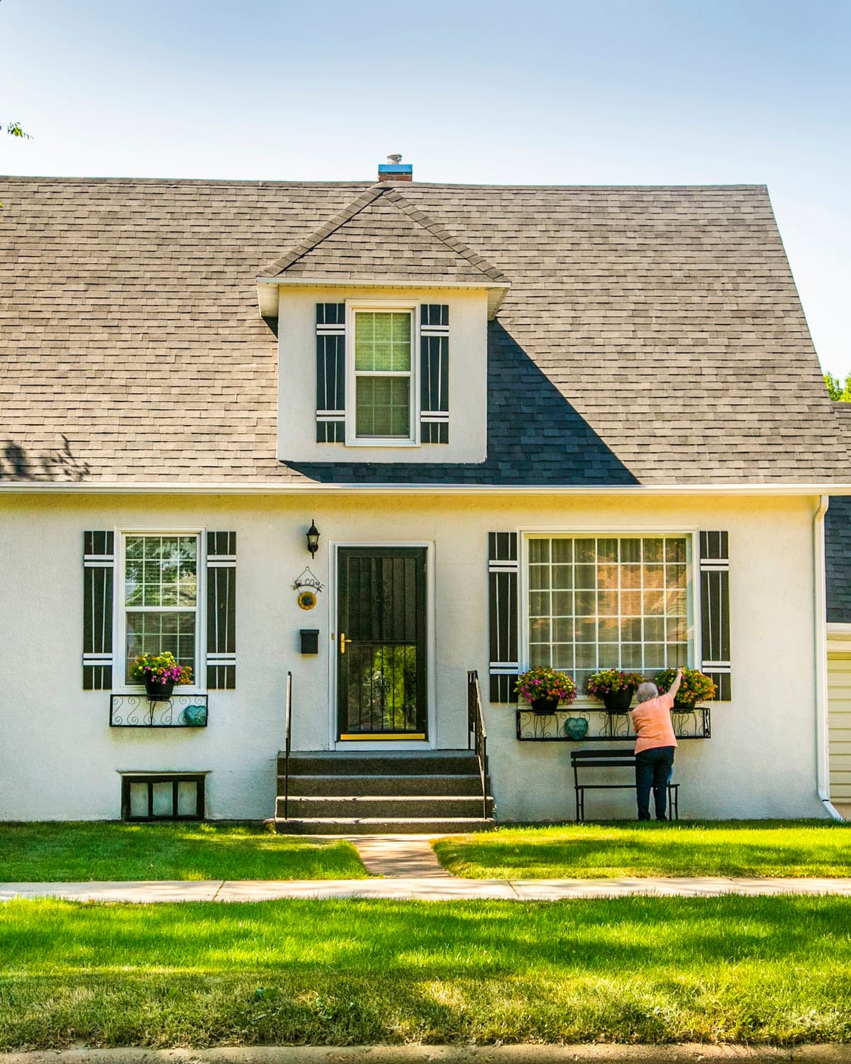White Cape Cod-style house with dark asphalt-shingle roof, attached garage and green front lawn under blue sky